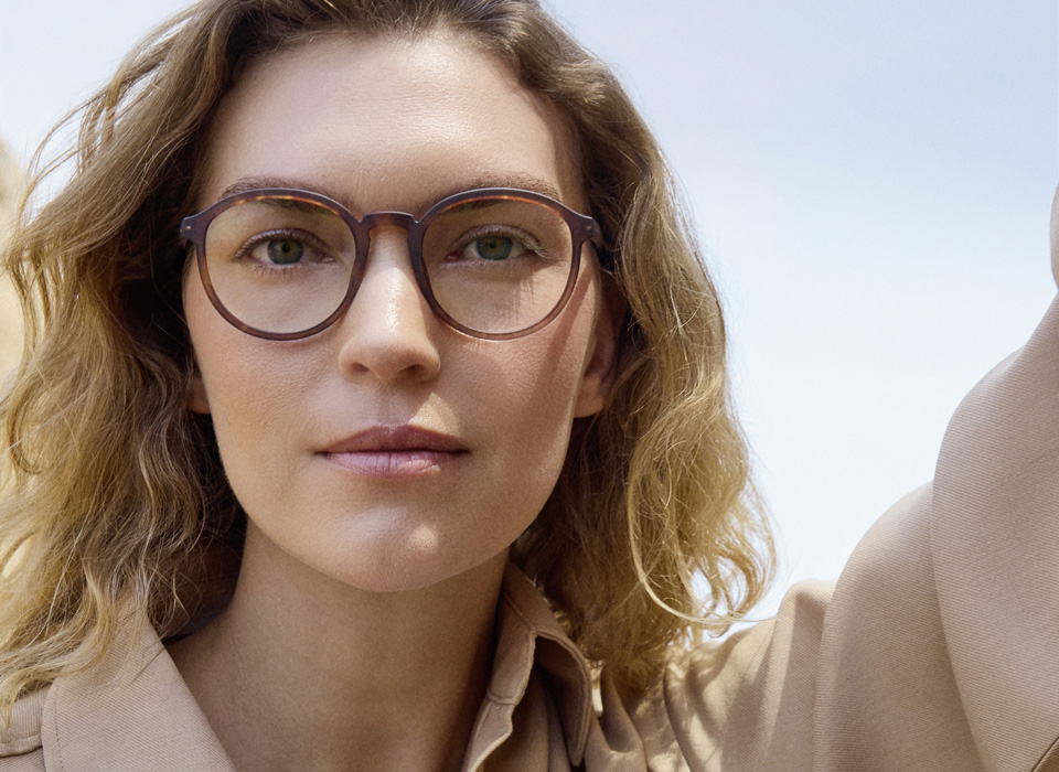 Arizona wears neutral colours and poses against a pale blue sky. She wears rich brown spectacles in a vintage style