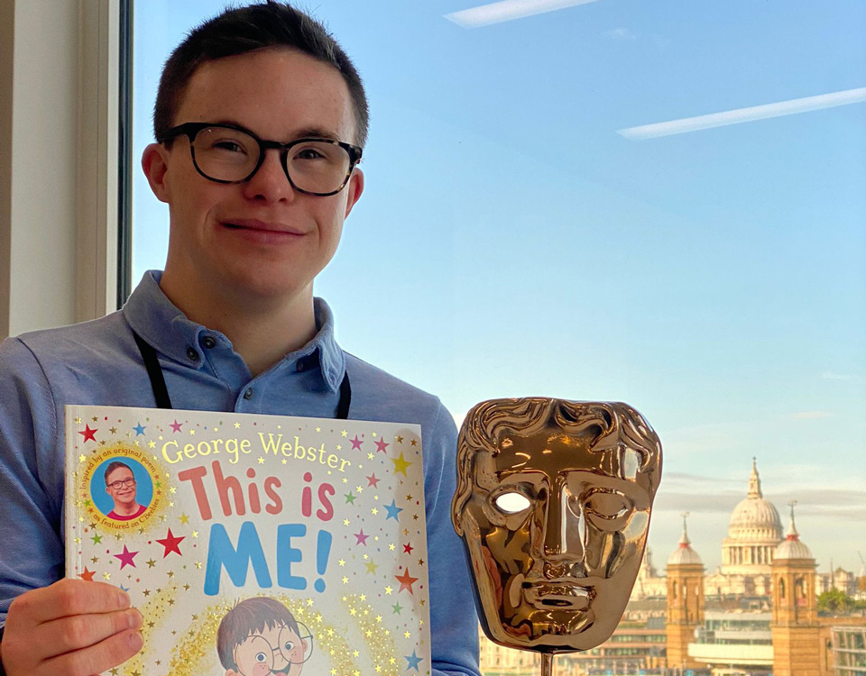 George sits at a desk in front of a window with a blue sky. He holds his book ‘This is Me’ and his BAFTA award – a golden mask on a blank square stand – is beside him