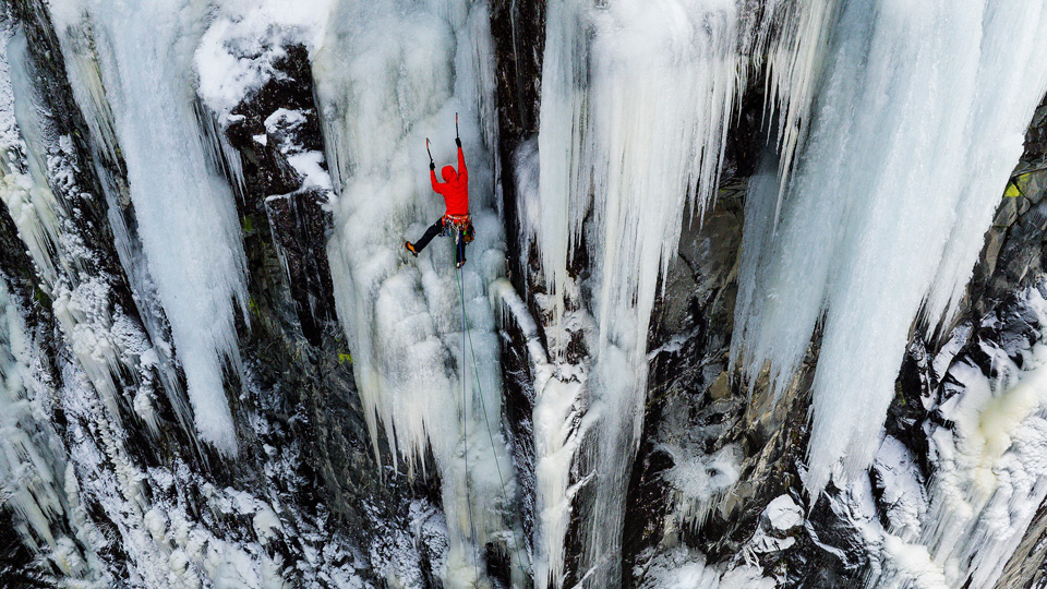 Wearing a bright red jacket, Tim is scaling a rockface covered in sheets of glass-like ice. He uses two picks or ice axes to pull himself up and lines trail from a belt of carabiners and climbing equipment at his waist. He is at the top of the frame, but the mountain face stretches down below him making him look small and very high up