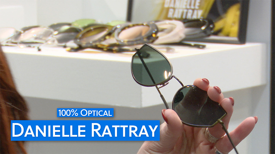 A pair of Danielle Rattray ‘Lowe’ sunglasses are held up in an exhibition stand – the frames have a antiqued metal look with blue tinted lenses. Blurred in the background are chunky acetate frames. 