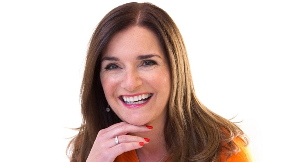 Deborah smiles brightly. She wears a bright orange top