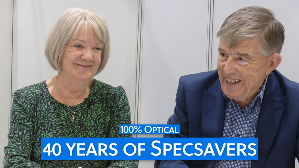 Dame Mary Perkins and Doug Perkins smile widely mid-interview. Mary wears a green and black printed dress and Doug is in a blue shirt and navy suit jacket
