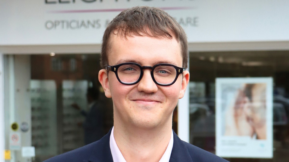 Jake stands outside of the Leightons Farnham practice. He wears black square glasses that are slightly rounded, with a navy blue suit jacket and pale pink shirt