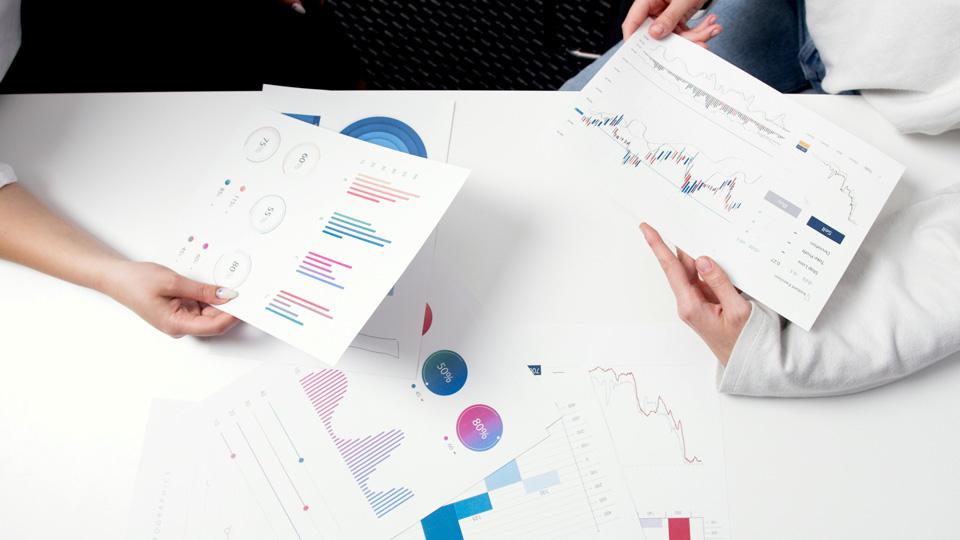 A bird’s eye view of an office table, where three people are holding out reports showing bar charts, pie charts and graphs