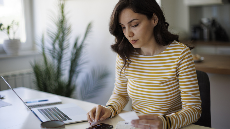 A woman with brown hair and a striped top sits at her laptop with some receipts