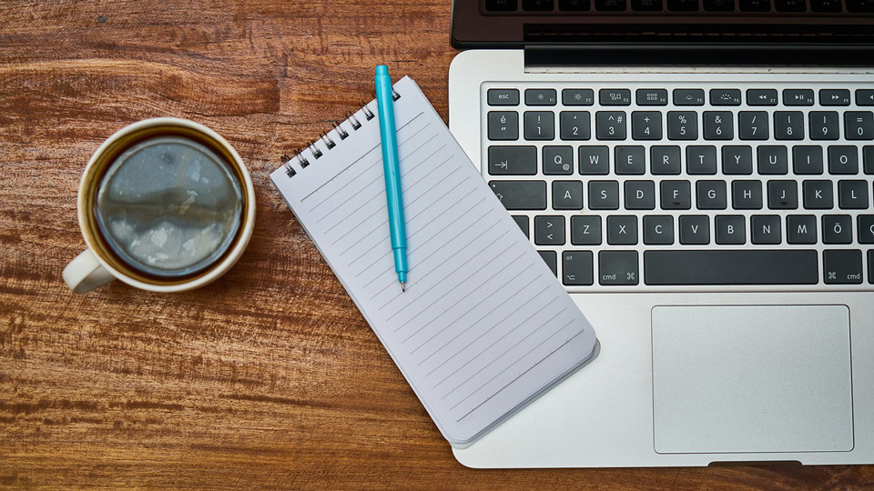 A desk shows an open laptop next to a mug of black coffee and blank spiral notebook