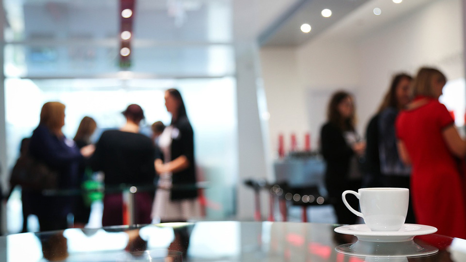 A conference room, the image is focused on a mug in the foreground while professionals network in the background 