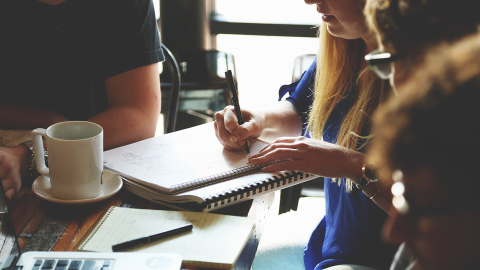 A close-up of a group of people gathered around a table with notebooks and a laptop. One lady is writing 