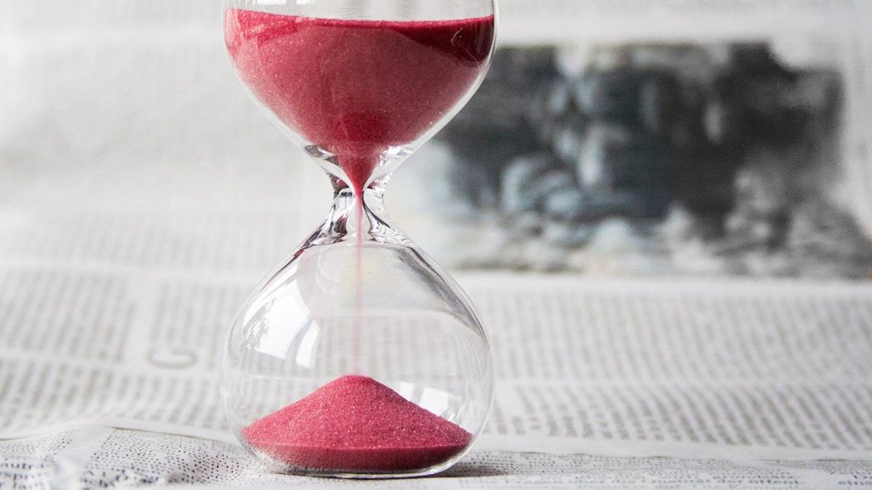 A sandtimer rests on a newspaper. Red sand is flowing from the full top to a growing pile in the bottom half