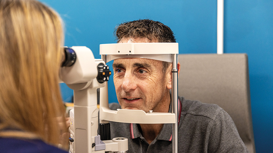 A female optometrist with long blonde hair is faced away from the camera. She examines the eye of a male patient with brown hair wearing a grey polo neck t shirt