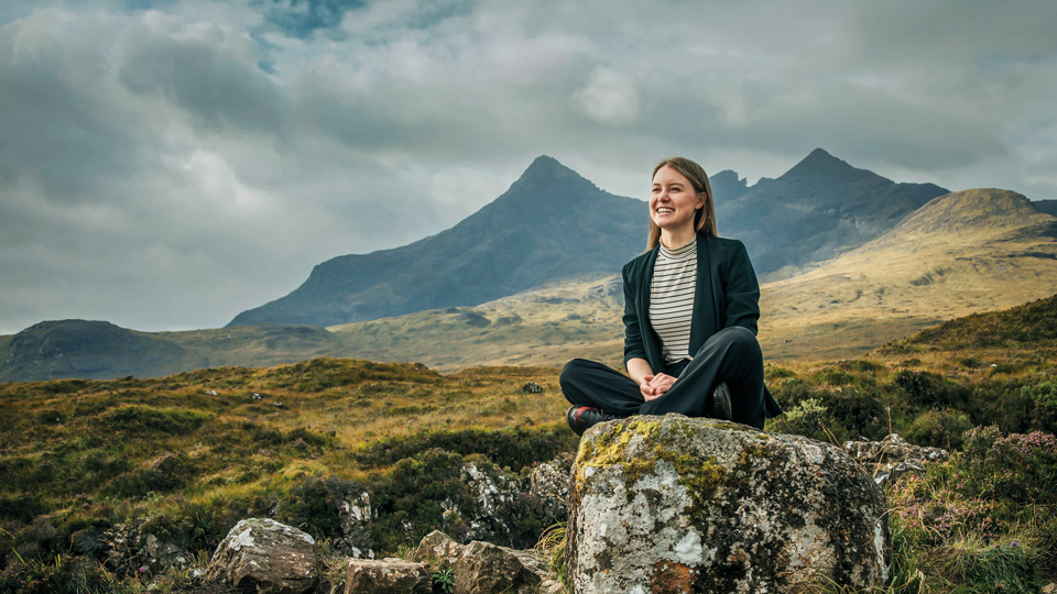 Optometrist Hannah Einarson is photographed on a rock with mountains in the background on the Isle of Skye