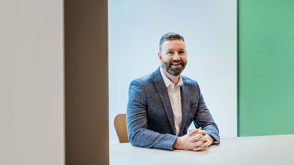 A man with a beard in a blue blazer and a white shirt sits at a grey table. His hands are clasped resting on the table. The wall behind him is grey and to the man’s left, the wall is green 