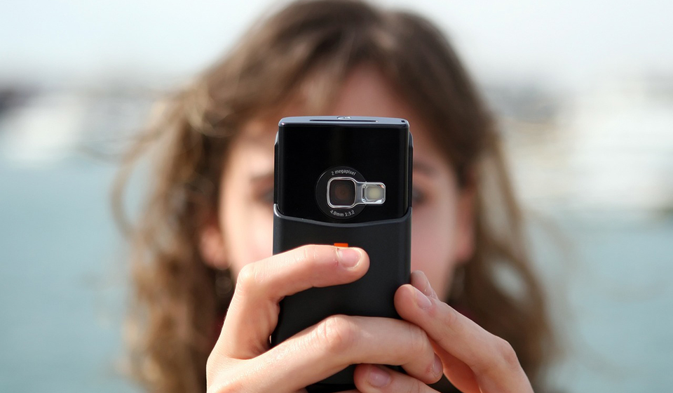 A woman holds a cellphone with both hands in front of her face. Her face and brown hair are out of focus in the background. 