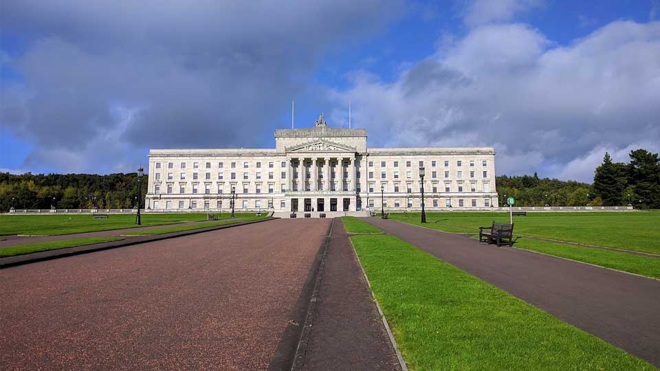 A road surrounded by neatly mown grass leads to a large stone-fronted government building with columns
