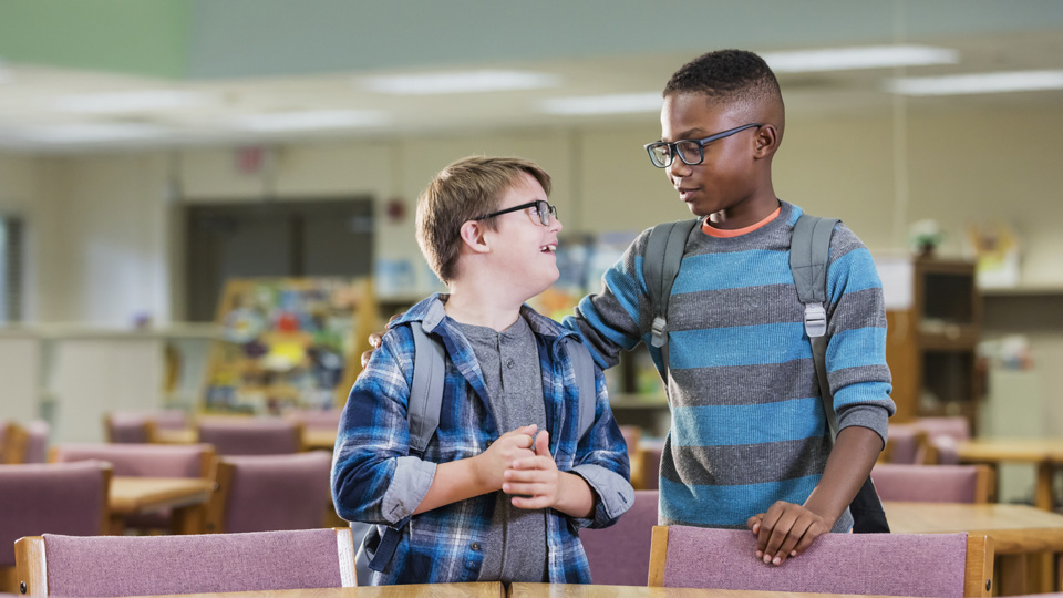 An 11-year-old Caucasian boy with down syndrome standing in the school library with his friend of the same age. Both are wearing glasses and are smiling at each other 