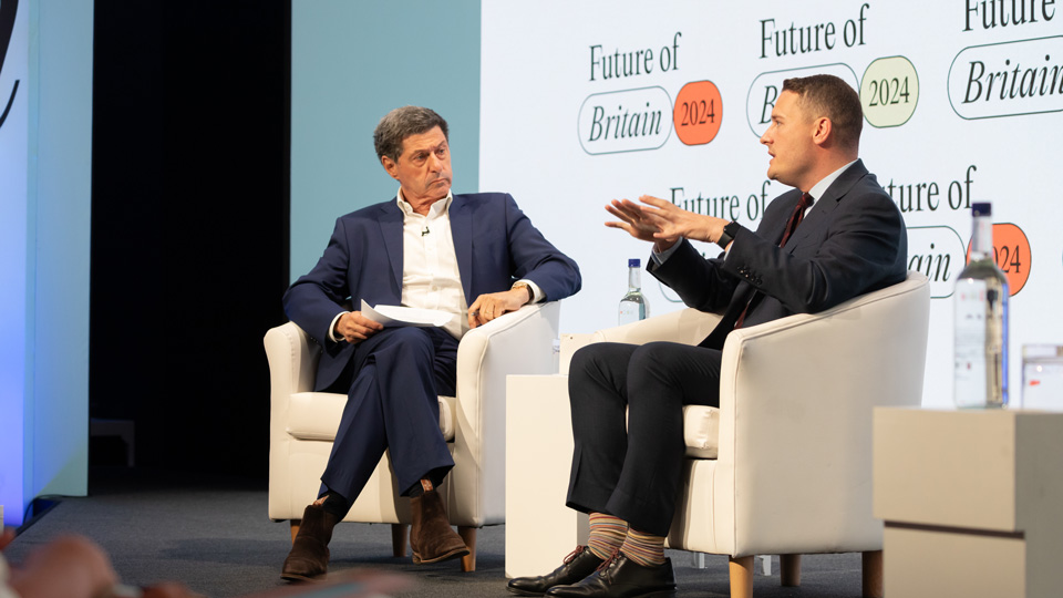 Two men are sat on cream chair on stage. Both are wearing dark suits and one is gesticulating with his hands 