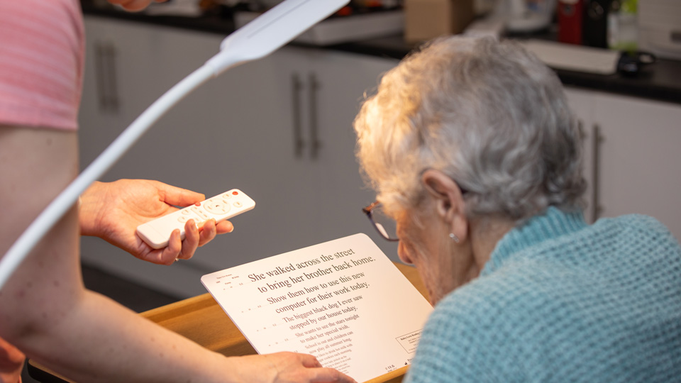 An older woman in an aquamarine jersey sits with her back to the camera at a wooden desk and light. She is reading from a tablet that is elevated from the table with a stand. On her left, another woman stands by the desk, pointing at the text with one hand and holding a remote control in the other