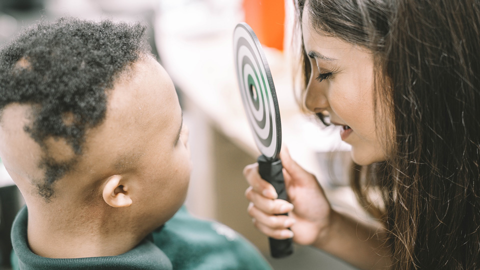 Optometrist Janki Vadgama leans forward and performs an eye test on a mixed-race male student with special needs 