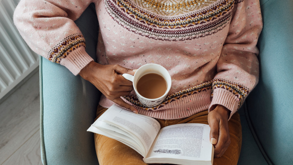 A person in a pink jumper and mustard cord trousers is reading a book and holding a cup of tea  