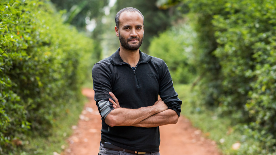 Dr Andrew Bastawrous, a man standing with his arms crossed on a country path with high bushes either side. 