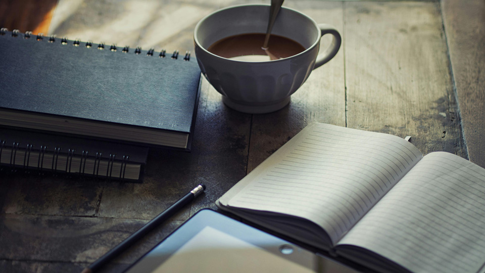 A scene of a table with notebooks piled up, a tablet, and a steaming hot drink 