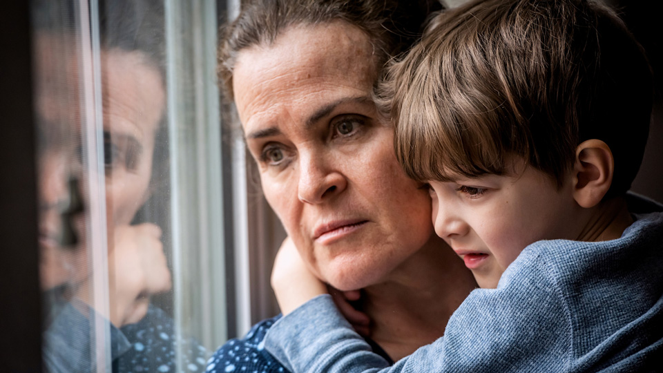Pensive woman with brown hair posing with her son, looking very concerned as she looks through a window 
