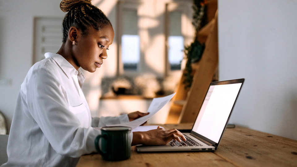  A black woman with her hair in braids is holding a piece of paper and frowning at a laptop screen  