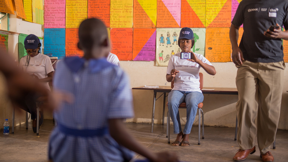 A vision screening takes place in a school in Zimbabwe, a little girl facing away from the camera towards a screener. The room is bright and colourful 