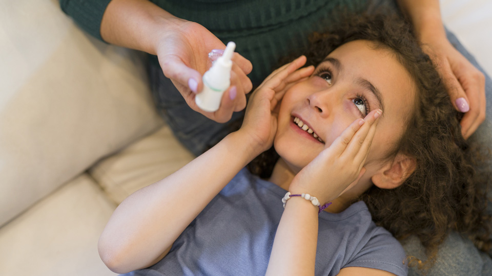 A woman sitting on a couch in a green top instils eye drops in a girl’s eyes.