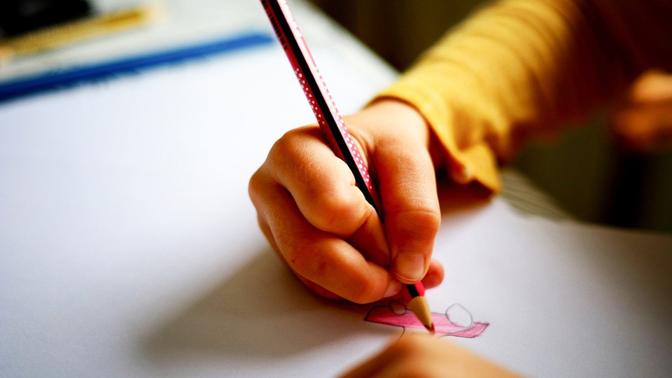 A child wearing a yellow long-sleeved top draws a pink car using a red pencil. 