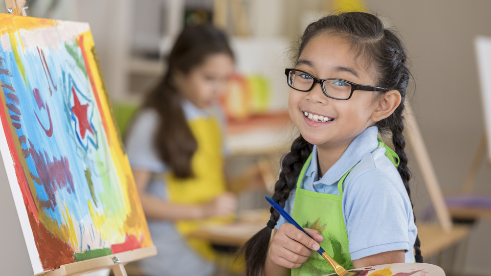 A young girl wearing glasses paints at an easel