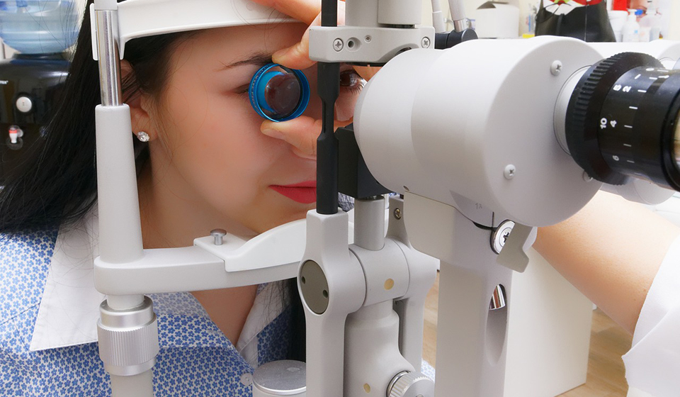 A woman wearing a blue patterned shirt has her eye examined. She rests her chin on white optical equipment while an optometrist, who is out of shot, places a lens close to her eye. 