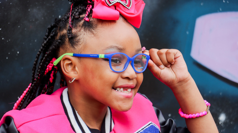 A young girl wearing a bright pink sport jacket and bow in her hair wears spectacles in a blue and green