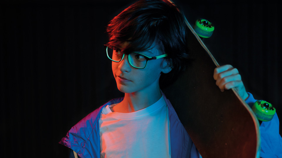 A young boy wearing frames which have glowing green details, holds a skateboard with green wheels
