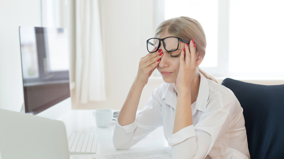 A young woman in a white shirt and glasses runs her eyes whilst sat in front of a computer screen 