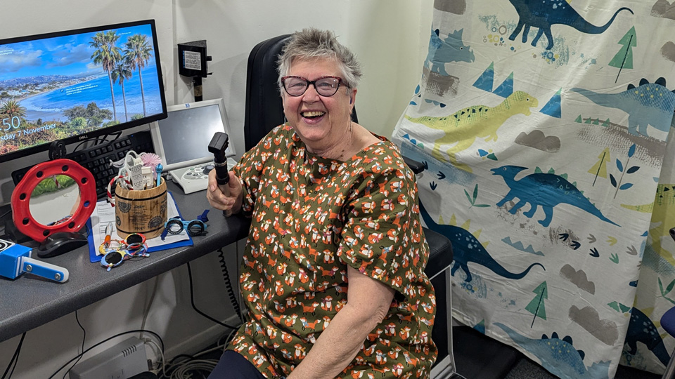 Professor Maggie Woodhouse sits at a desk in a clinic room with a bright smile. Her desk has a barrel filled with props, next to a light-up mirror, several pairs of children’s frames