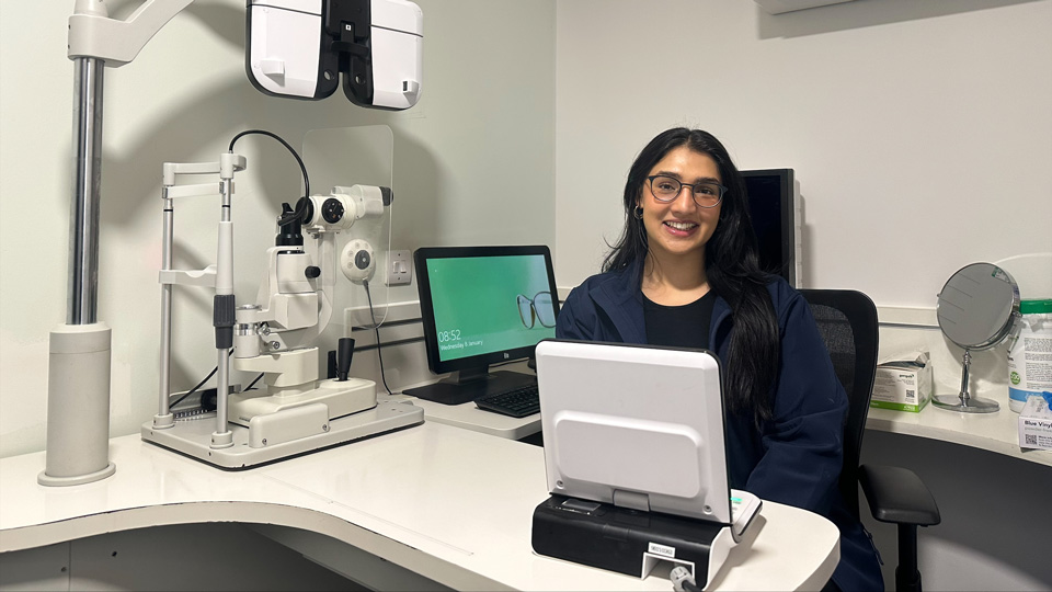 Samiriah sits behind her desk in the examination room of an optometry practice. Around her is testing equipment and a computer monitor