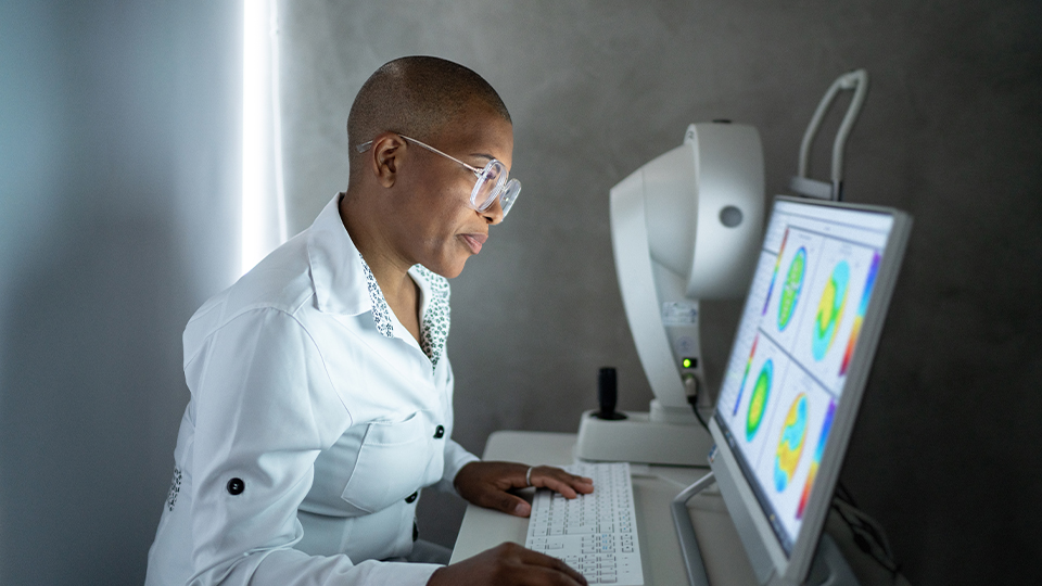 A female clinician in a white shirt examines images on a computer screen with sight testing equipment in the background 