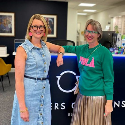 Two women stand in front of the reception desk of Peters Opticians. Ghislaine stands on the left, wearing a denim dress, thick framed spectacles and bright pink circle earrings. Elohdi stands on the right in a green and pink sweatshirt with clear-green spectacles.