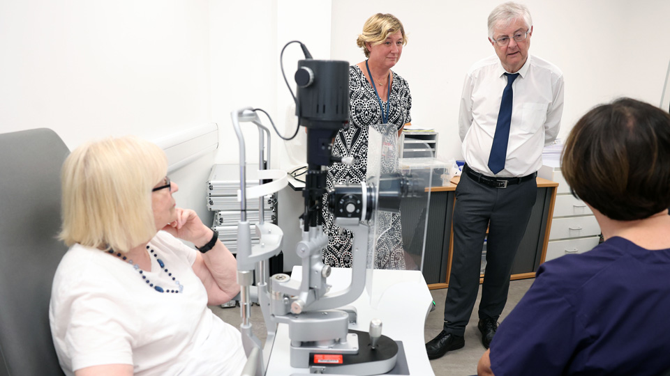 Mark Drakeford appears in conversation with a practitioner who is sat at a slit lamp with a patient. The practitioner has their back to the camera as they speak with the First Minister. Professor Barbara Ryan stands to his left.    