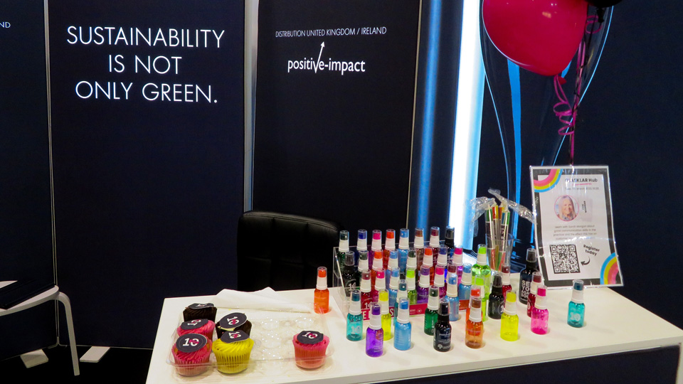 An exhibition stand has an array of rainbow-hued solution bottles, red balloons and cupcakes. Behind the table, banners showcase the colourful bottles with the words ‘Sustainability is not only green’.